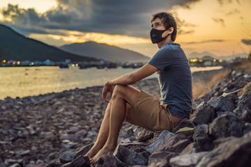 Fototapeta na wymiar Self-isolation in nature. Young man in Fashionable black medical mask with filter sitting by the river. Coronavirus 2019-ncov epidemic concept. Man in a black medical mask. Portrait of a man with