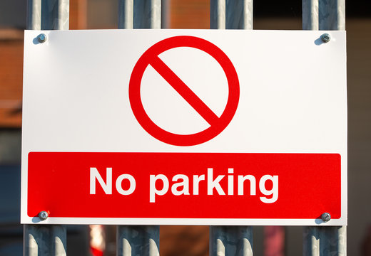 Close up view of a red & white No Parking sign attached to a metal security fence, at an industrial trading estate in Colliers Wood, London, UK.