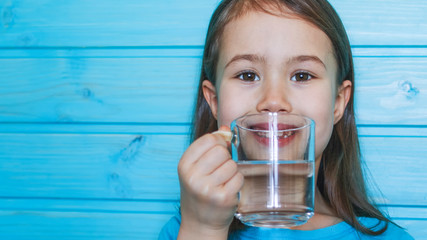 Beautiful little girl drinks clean water from a transparent glass on a blue background