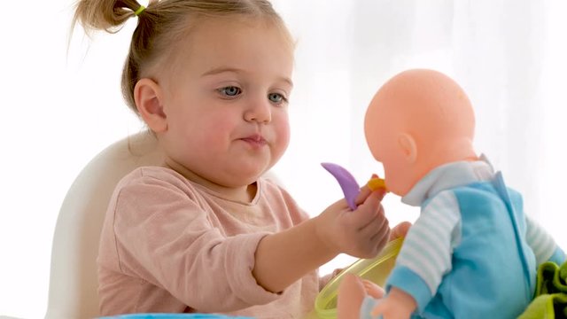 Funny little girl puffing cheeks and giving cereals to baby doll while having breakfast at home in morning