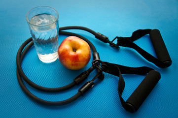 Sport expander, glass with water and an apple on blue yoga mat background