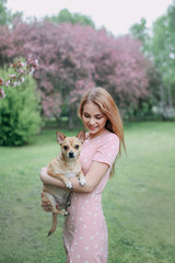 Beautiful gentle dreamy sweet pretty happy girl in a delicate dress and hat walking with her pet, with a dog in the park among the blooming apple trees in spring, friendship