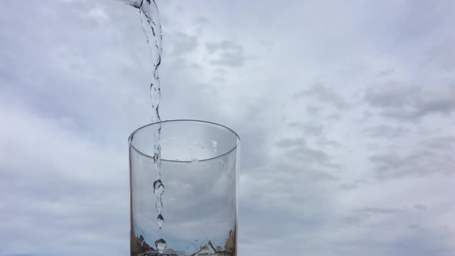 Llenando un vaso de agua con una jarra a c&aacute;mara lenta con un cielo de nubes al fondo.
