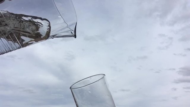 Llenando un vaso de agua con una jarra a c&aacute;mara lenta con un cielo de nubes al fondo.