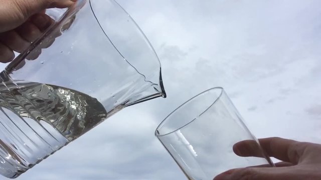 Llenando un vaso de agua con una jarra a c&aacute;mara lenta con un cielo de nubes al fondo.