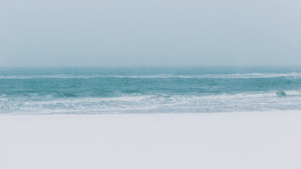 Winter sea at noon. On the beach lies snow, a cold turquoise sea. Slight fog over the sea and gray sky