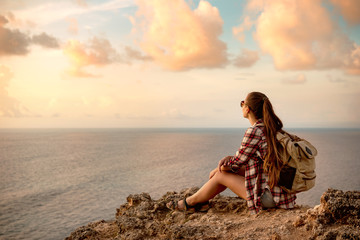 Girl with backpack sits on the cliff against sea