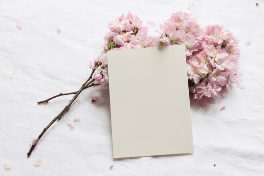 Wedding spring styled stock photo. Feminine desktop mockup scene with pink blossoming Japanese cherry tree branches and blank paper greeting card on white linen table background. Flat lay, top view.