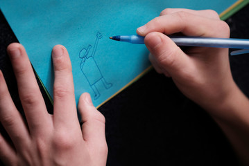 Hands of a teenager close-up draws a cartoon from a simple person on blue paper on a black background with empty space for text