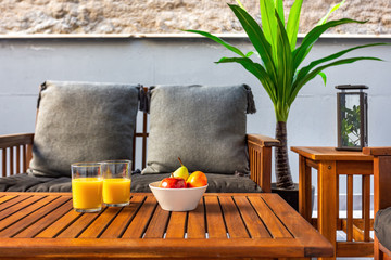 Terrace with wooden table, sofa and chairs. Fruits and orange juice at the table. Having healthy breakfast at home. Vacation and travel concept. Bed and breakfast.