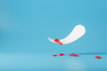 Sanitary pad in flight on a blue background with fallen petals of red flowers. Concept of the beginning of menopause.