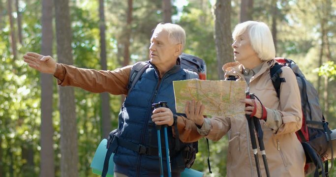 Caucasian old married couple standing in forest and examining route plan. Tourists looking at map and choosing path while hiking in wood. Senior man and woman hikers outdoor. Tourism concept.