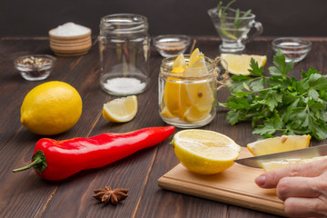 Hands chop a lemon on cutting board. Lemon in jar. Chili pepper, whole lemon and salt.