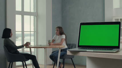 Multiracial girlfriends talking while sitting at the table on kitchen and drinks tea, laptop with green screen on foreground. 