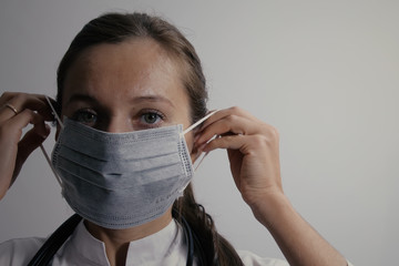 Close-up portrait of young adult female doctor put a medical mask on her face and looking at camera. Physician in protective mask