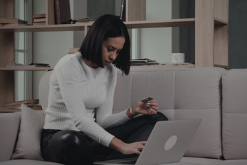 African American female with straith short hair in casual clothes seating on couch and entering credit card credentials on laptop while shopping online at home