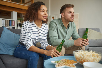 Obraz na płótnie Canvas Warm-toned portrait of modern mixed-race couple watching TV at home and drinking beer while sitting on sofa in cozy apartment, copy space