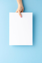 Young woman’s hand holding blank paper sheet on blue background. Mockup