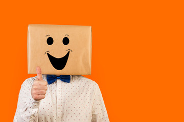 Hombre con caja de cartón en la cabeza con gesto positivo y feliz y el dedo pulgar de la mano hacia arriba OK sobre fondo naranja liso y aislado. Vista de frente y de cerca. Copy space