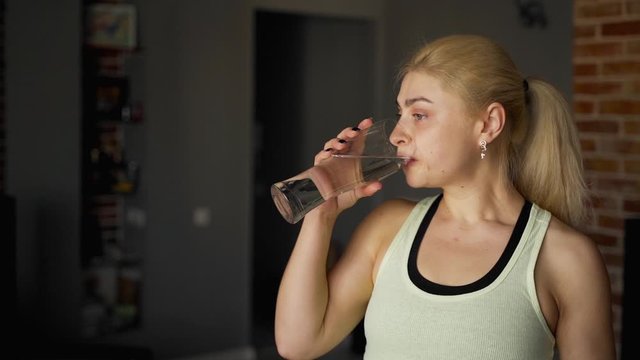 Trainings at home: A strong beautiful fitness girl takes a glass of water and drik after training. Woman stay at her living room. She in athletic sport wear. Slow motion