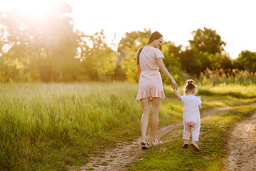 Fototapeta premium Mother and little daughter playing together in a summer park at sunset. Happy mom with small child having fun on sunny field. Kisses and hugs. The concept of a happy family. 
