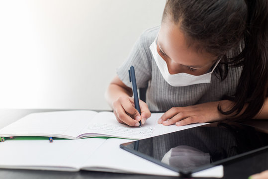 Little girl wears a prevention mask doing her homework during the quarantine. Concept of studying during coronavirus covid19 infection.