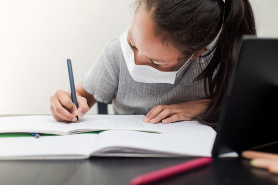 Little girl in a protective face mask does homework for school during quarantine. Distance learning online education during the pandemic of coronavirus Covid-19.