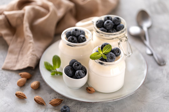 Non-dairy alternative. Vegetable yogurt in jars on a white background. Almond cream in a glass jar and fresh blueberry on a light plate and almond seeds. Healthy eating concept.