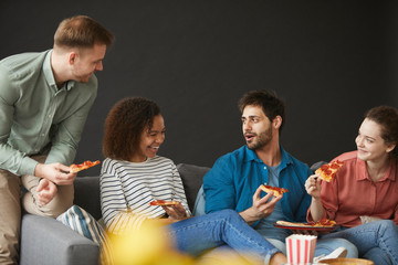 Multi-ethnic group of friends eating pizza and snacks while enjoying party at home sitting on big sofa against black background, copy space above