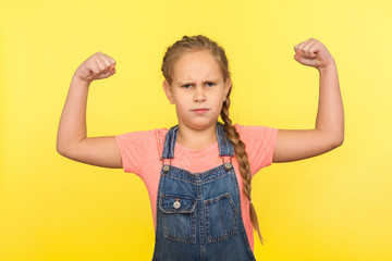 I'm strong! Portrait of brave little girl with braid in denim overalls raising hands showing...