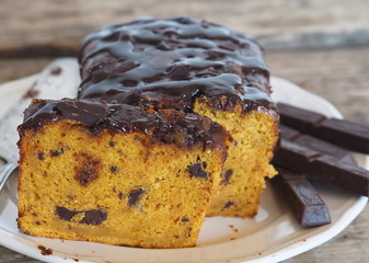 Festive pumpkin chocolate soft muffin, poured with chocolate icing with cut pieces on a white plate on a wooden background.