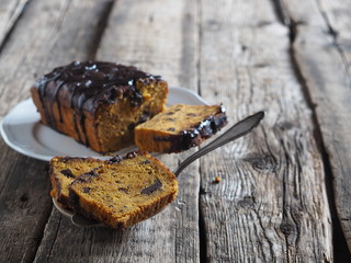 Festive pumpkin chocolate soft muffin, poured with chocolate icing with cut pieces on a white plate on a wooden background.