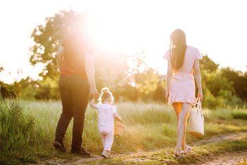 Fototapeta premium Happy young family walking in the park at sunset. Mom, dad and little daughter having fun in summer park. The concept of a happy family. Parents hold the baby's hands. Kisses and hugs.