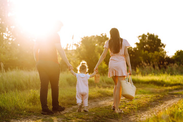 Naklejka na ściany i meble Happy young family walking in the park at sunset. Mom, dad and little daughter having fun in summer park. The concept of a happy family. Parents hold the baby's hands. Kisses and hugs.