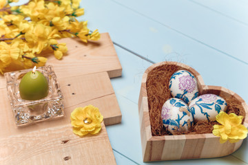 Heart shaped box with decorated Easter eggs, yellow flowers, burning candle and brown boards on wooden blue background. Spring and Easter concept. Close up, copy space.