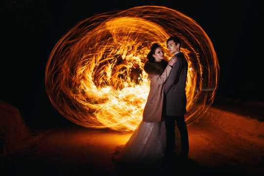 The groom embraces the bride against the background of flames, standing in the snow. Technique of night photography by freezing light and lightpainting.