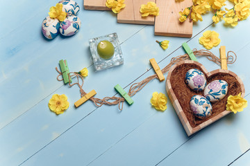 Heart shaped box with decorated Easter eggs, yellow flowers, burning candle and brown boards on wooden blue background. Spring and Easter concept. Top view, flatlay, copy space.