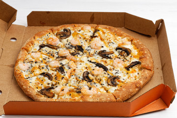 Closeup pizza with mushrooms and shrimp in open cardboard box on white wooden table. Shallow focus.