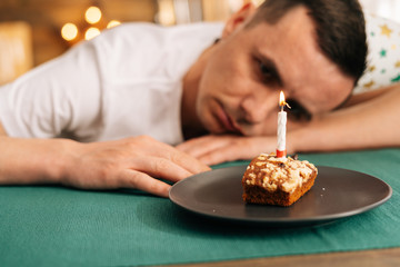Sad lonely young man in festive hat celebrating birthday alone, sitting at the birthday cake and...
