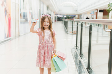 Cute little girl in a pink dress holds a credit card for shopping in a shopping center. A beautiful child in the mall makes shopping. Online Shopping Concept