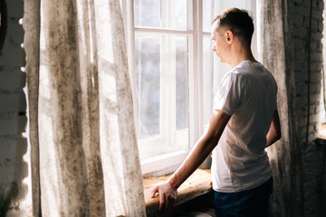 Thoughtful European young man stands by the window on sunny day, enjoying beautiful day.