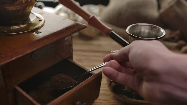 The unrecognizable person taking ground coffee from an old coffee grinder to brassy Cezve. The traditional way of making ground coffee from roasted beans. Mechanical wooden authentic coffee grinder.