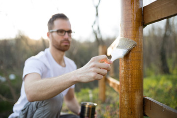 Unrecognizable young man painting wooden fence in backyard, selective focus on a hand holding the paintbrush in the foreground