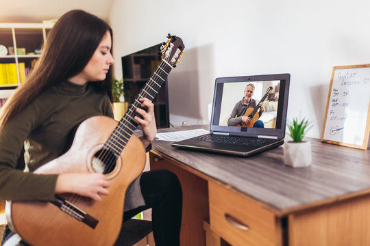 Focused girl playing acoustic guitar and watching online course on laptop while practicing at home. Online training, online classes.