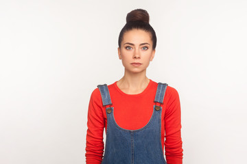 Portrait of stylish slim pretty girl with hair bun in denim overalls standing, looking at camera with serious face, calm pensive focused expression. indoor studio shot isolated on white background
