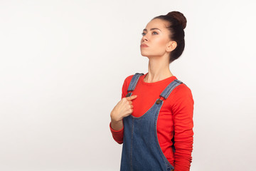 Portrait of arrogant egoistic girl with hair bun in denim overalls pointing herself and looking up...