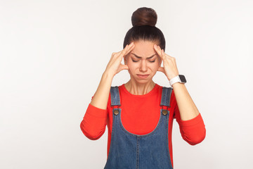 Headache. Portrait of tired depressed upset girl with hair bun in denim overalls feeling unhealthy, suffering migraine and tension, meningitis, flu symptom. studio shot isolated on white background