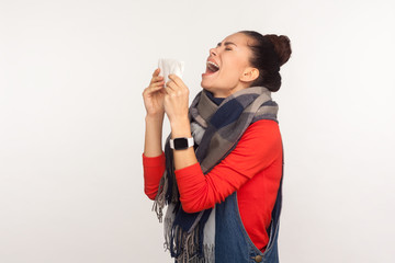 Flu symptoms. Portrait of unhealthy young woman wrapped in warm scarf sneezing hard in tissue, coughing feeling unwell, suffering influenza disease. indoor studio shoot isolated on white background