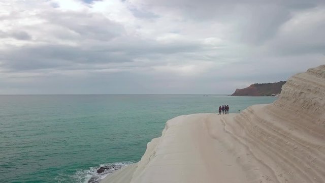 Aerial footage boom out and pan left of group of friends admiring the views from the white rock called Scala dei Turchi, Stairs of the Turkmen in Sicily, Italy. Cloudy day, turquoise water.