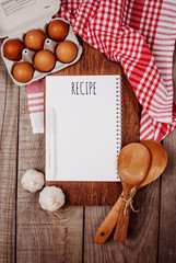 Wooden cutting board on a wooden background with garlic, ladles, eggs and a blank notebook with quote Recipe